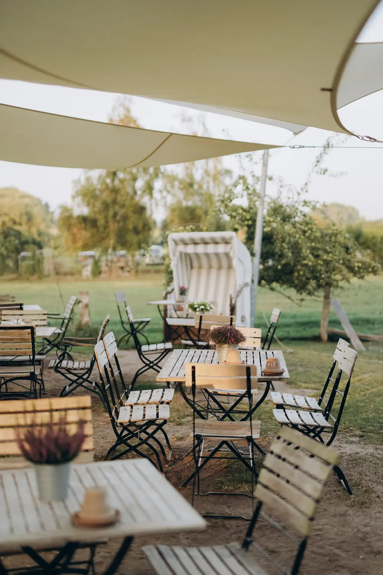 Gartenterrasse des Cafés mit Klappstühlen und Tischen unter weißen Sonnensegeln, im Hintergrund ein Strandkorb und Obstbäume