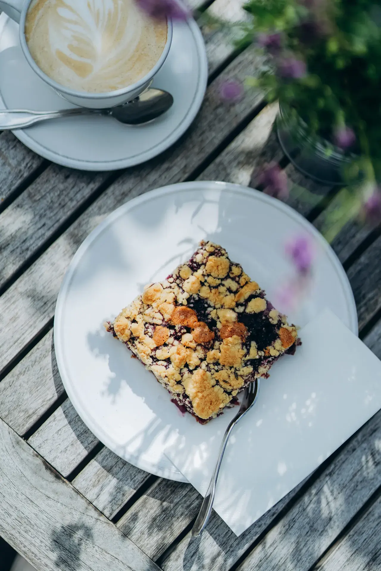 Stück Streuselkuchen mit Beeren auf weißem Teller mit Gabel, daneben ein Cappuccino mit Milchschaum-Muster